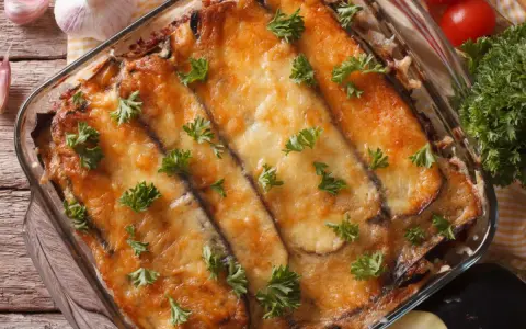 Ein Moussaka, ein Klassiker der griechischen Rezepte, in einer Auflaufform, mit geschmolzenem Käse und Petersilie garniert. Daneben liegen Knoblauch und Tomaten.