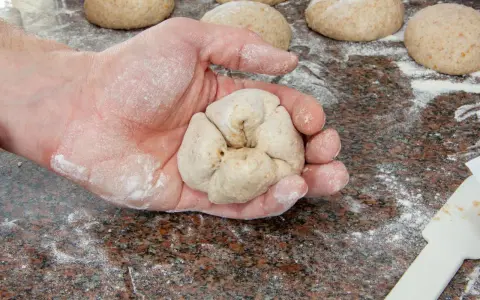 Eine Hand formt Teiglinge für das Backen mit Vollkornmehl. Mehrere Teiglinge liegen bereit auf einer bemehlten Arbeitsfläche.