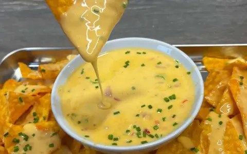 Ein Tortilla-Chip taucht in eine Schüssel mit cremiger Chili Cheese Sauce, garniert mit Schnittlauch.