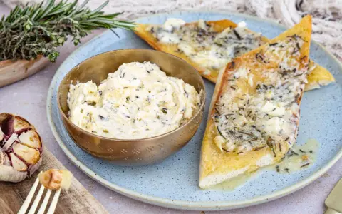 Schüssel mit selbstgemachter Knoblauchbutter, daneben gerösteter Knoblauch und Kräuterbrot. Perfekt zum **knoblauchbutter selber machen**.