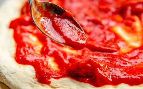 Eine Löffelspitze verteilt die leuchtend rote Pizza Sauce wie in der Pizzeria auf den Teig.