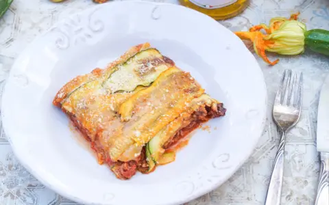 Ein Stück Zucchini-Lasagne mit Hackfleisch und Tomatensauce, bestreut mit Parmesan. Daneben liegen frische Zucchini und eine Blüte.
