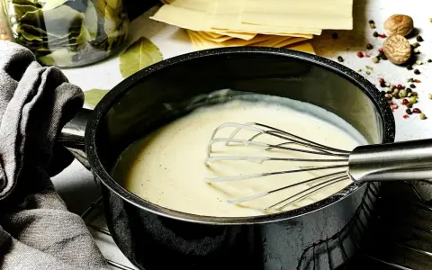 Cremige Bechamelsauce in einem Topf, bereit für die Lasagne. Daneben liegen Lasagneplatten, Lorbeerblätter und Gewürze.