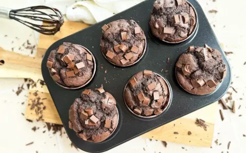 Sechs saftige Muffins mit Schokoladenstückchen in einer Muffinform. Ein Schneebesen mit Schokoladenspuren liegt daneben.