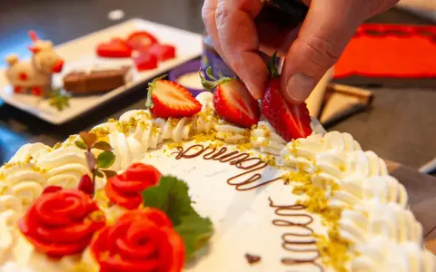 Kreative torten verzieren ideen: Eine Hand platziert Erdbeeren auf einer Sahnetorte mit Schokoladenschrift und roten Rosen.