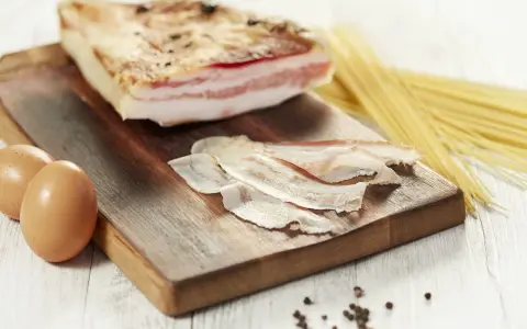 Zutaten für Guanciale Carbonara: ein Stück Guanciale, Eier, Spaghetti und Pfefferkörner auf einem Holzbrett.