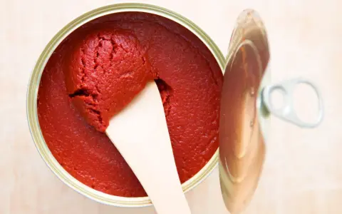 Ein offener Behälter mit leuchtend rotem Tomatenmark, aus dem mit einem Holzspatel eine Portion entnommen wird. Perfekt als Tomatenmark-Ersatz für Saucen und Eintöpfe.