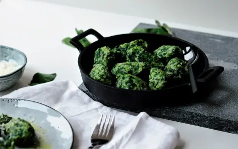Grüne Malfatti, zubereitet nach einem einfachen Rezept, serviert in einer Gusseisenpfanne. Daneben ein Schälchen mit geriebenem Käse und ein Teller mit den fertigen Malfatti.