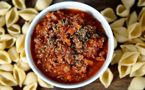 Herzhaftes Ragout alla Bolognese in einer Schüssel, umgeben von ungekochten Muschelnudeln.
