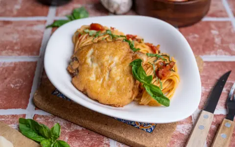 Ein Teller Spaghetti mit Tomatensauce und einem panierten Schnitzel, das ist Piccata. Frisch garniert mit Basilikum.