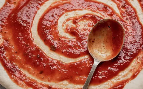 Ein Löffel verteilt das rote Pizzasaucen-Rezept auf einem Teigboden in Spiralform.