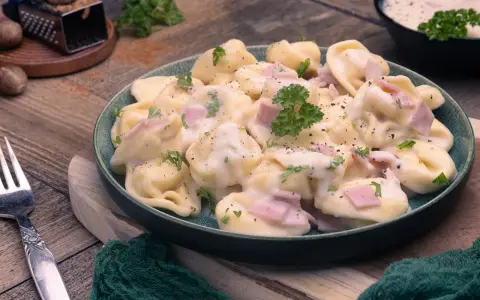 Ein Teller mit cremigen Tortellini alla Panna, garniert mit Petersilie und Schinkenwürfeln. Dazu eine Gabel und Muskatnuss.