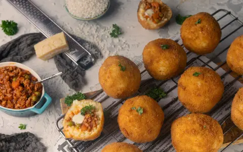 Goldbraune Arancini, gefüllt mit Ragout, liegen auf einem Rost. Zutaten für das Arancini Rezept wie Reis, Parmesan und Hackfleisch sind daneben arrangiert.