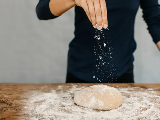 Eine Hand streut Mehl über einen Teig auf einem Holztisch. Das ist der Beginn vom backen mit glutenfreiem Mehl.