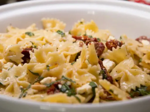Farfalle-Nudelsalat mit getrockneten Tomaten, Feta und Pinienkernen, garniert mit frischen Kräutern. Ein köstliches Gericht.