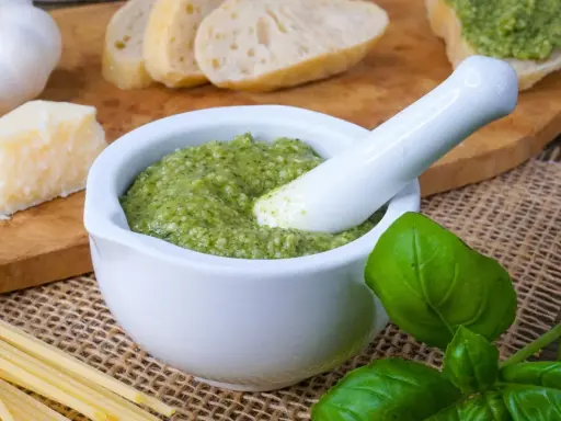 Frisches Pesto im Mörser, bereit für ein köstliches Rezept. Daneben liegen Brot, Parmesan, Knoblauch und Basilikum.
