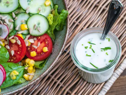 Frischer Salat mit Tomaten, Gurken und Mais, daneben ein Glas mit selbstgemachtem Joghurt-Dressing, garniert mit Schnittlauch.