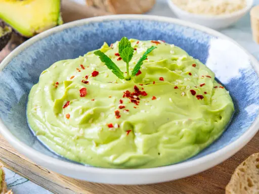 Cremige Avocado-Creme in einer blauen Schüssel, garniert mit Minze, Sesam und Chiliflocken. Daneben liegen Brotscheiben.