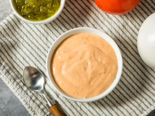 Cremige, orangefarbene Big Mac Sauce in einer Schüssel, daneben gehackte Gurken, eine Tomate und eine Zwiebel.