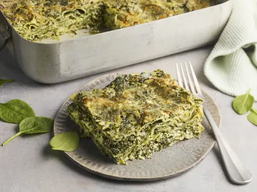 Ein Stück Spinatlasagne auf einem Teller, daneben frische Spinatblätter und eine Gabel. Das Spinatlasagne rezept ist ein Genuss!