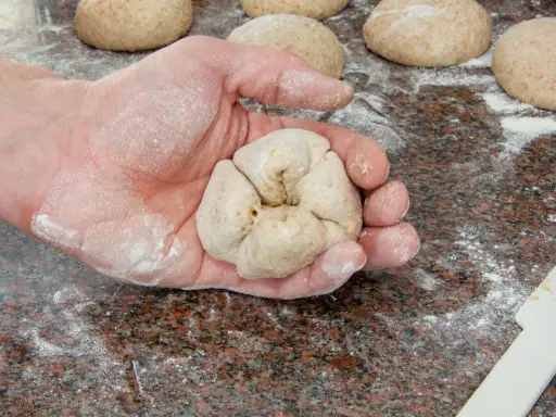 Eine Hand formt Teiglinge für das Backen mit Vollkornmehl. Mehrere Teiglinge liegen bereit auf einer bemehlten Arbeitsfläche.
