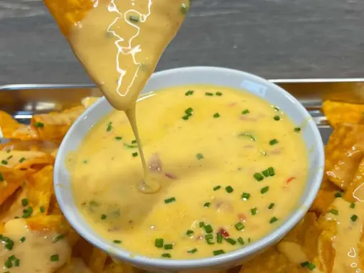 Ein Tortilla-Chip taucht in eine Schüssel mit cremiger Chili Cheese Sauce, garniert mit Schnittlauch.