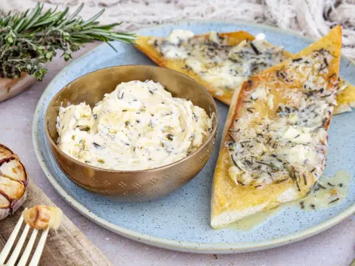 Schüssel mit selbstgemachter Knoblauchbutter, daneben gerösteter Knoblauch und Kräuterbrot. Perfekt zum **knoblauchbutter selber machen**.