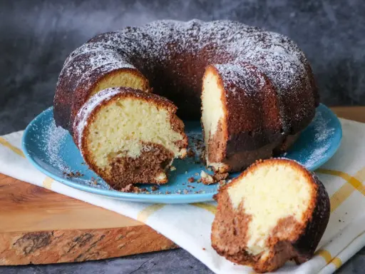 Saftiger weltbester Marmorkuchen, mit Puderzucker bestäubt, auf blauem Teller, ein Stück abgetrennt.