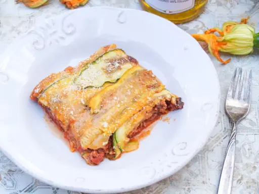 Ein Stück Zucchini-Lasagne mit Hackfleisch und Tomatensauce, bestreut mit Parmesan. Daneben liegen frische Zucchini und eine Blüte.
