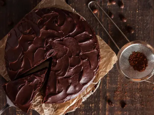 Ein saftiger Nutella Kuchen mit dunkler Schokoladenglasur, ein Stück wurde bereits entnommen. Daneben ein Sieb mit Kakaopulver.