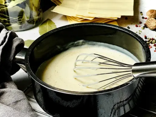 Cremige Bechamelsauce in einem Topf, bereit für die Lasagne. Daneben liegen Lasagneplatten, Lorbeerblätter und Gewürze.