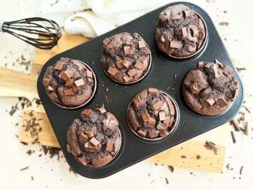 Sechs saftige Muffins mit Schokoladenstückchen in einer Muffinform. Ein Schneebesen mit Schokoladenspuren liegt daneben.