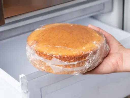 Hand nimmt einen in Frischhaltefolie eingewickelten Kuchen aus dem Kühlschrank, um ihn aufzutauen.