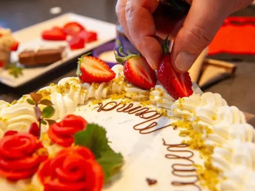 Kreative torten verzieren ideen: Eine Hand platziert Erdbeeren auf einer Sahnetorte mit Schokoladenschrift und roten Rosen.