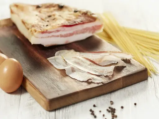 Zutaten für Guanciale Carbonara: ein Stück Guanciale, Eier, Spaghetti und Pfefferkörner auf einem Holzbrett.