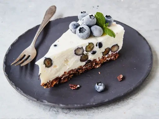 Ein Stück Heidelbeertorte ohne Backen auf schwarzer Platte, garniert mit frischen Heidelbeeren und Minze.