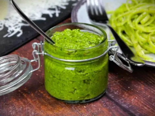 Frisches Rucola Pesto in einem Glas mit Löffel, daneben eine Portion Pasta.