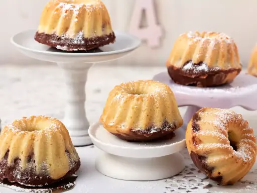 Mehrere kleine, mit Puderzucker bestäubte Mini Gugelhupf-Kuchen mit Schokoladenboden stehen auf verschiedenen Etageren.