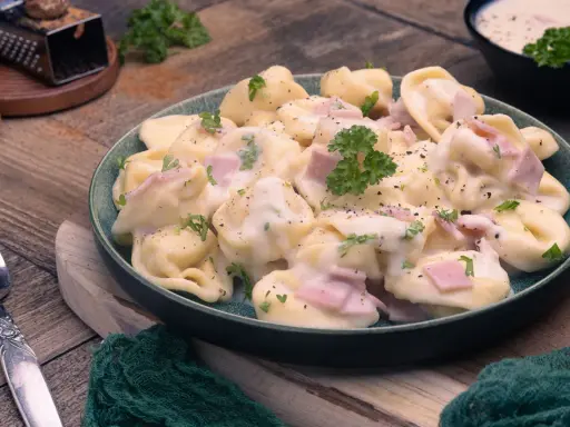 Ein Teller mit cremigen Tortellini alla Panna, garniert mit Petersilie und Schinkenwürfeln. Dazu eine Gabel und Muskatnuss.
