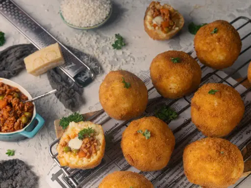 Goldbraune Arancini, gefüllt mit Ragout, liegen auf einem Rost. Zutaten für das Arancini Rezept wie Reis, Parmesan und Hackfleisch sind daneben arrangiert.