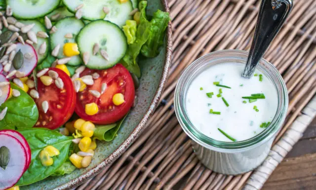 Frischer Salat mit Tomaten, Gurken und Mais, daneben ein Glas mit selbstgemachtem Joghurt-Dressing, garniert mit Schnittlauch.