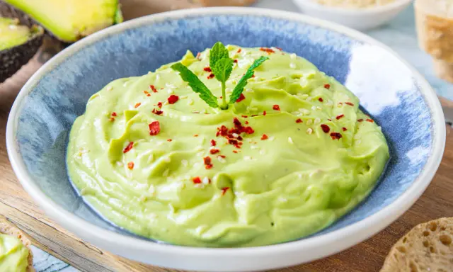Cremige Avocado-Creme in einer blauen Schüssel, garniert mit Minze, Sesam und Chiliflocken. Daneben liegen Brotscheiben.