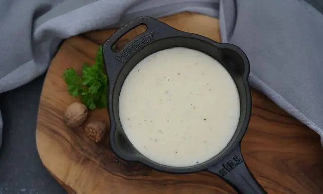 Cremige Béchamelsauce in einer kleinen Gusseisenpfanne, garniert mit Petersilie und Muskatnüssen.
