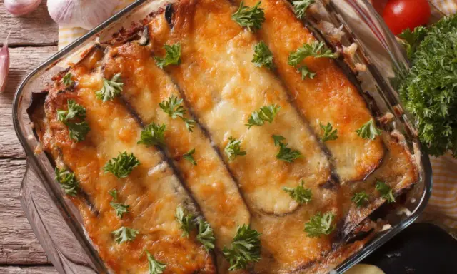 Ein Moussaka, ein Klassiker der griechischen Rezepte, in einer Auflaufform, mit geschmolzenem Käse und Petersilie garniert. Daneben liegen Knoblauch und Tomaten.