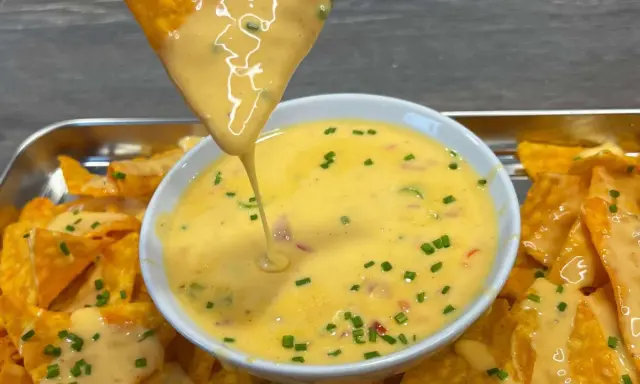 Ein Tortilla-Chip taucht in eine Schüssel mit cremiger Chili Cheese Sauce, garniert mit Schnittlauch.