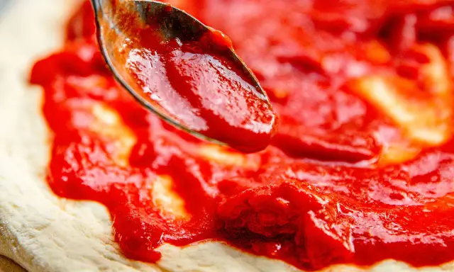 Eine Löffelspitze verteilt die leuchtend rote Pizza Sauce wie in der Pizzeria auf den Teig.