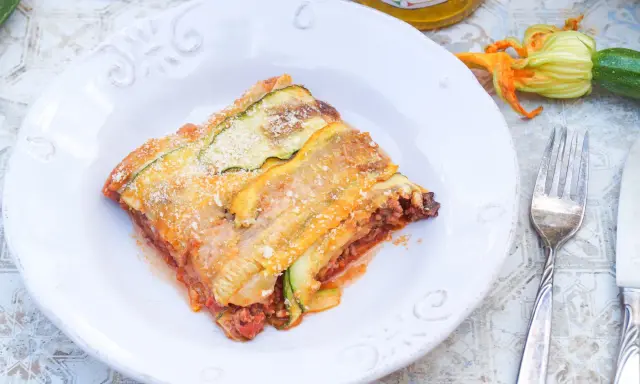 Ein Stück Zucchini-Lasagne mit Hackfleisch und Tomatensauce, bestreut mit Parmesan. Daneben liegen frische Zucchini und eine Blüte.