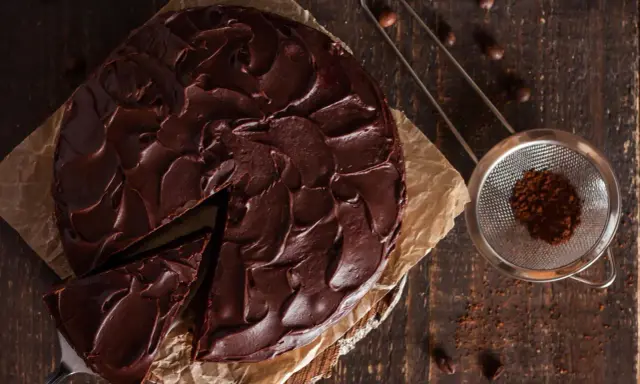 Ein saftiger Nutella Kuchen mit dunkler Schokoladenglasur, ein Stück wurde bereits entnommen. Daneben ein Sieb mit Kakaopulver.