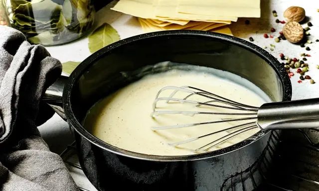 Cremige Bechamelsauce in einem Topf, bereit für die Lasagne. Daneben liegen Lasagneplatten, Lorbeerblätter und Gewürze.