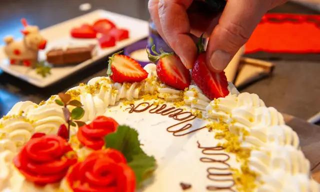 Kreative torten verzieren ideen: Eine Hand platziert Erdbeeren auf einer Sahnetorte mit Schokoladenschrift und roten Rosen.